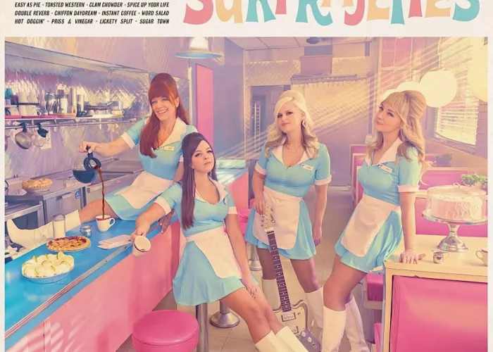 Four members of the Sufrajettes pose in a pink and chrome diner, wearing light blue uniforms