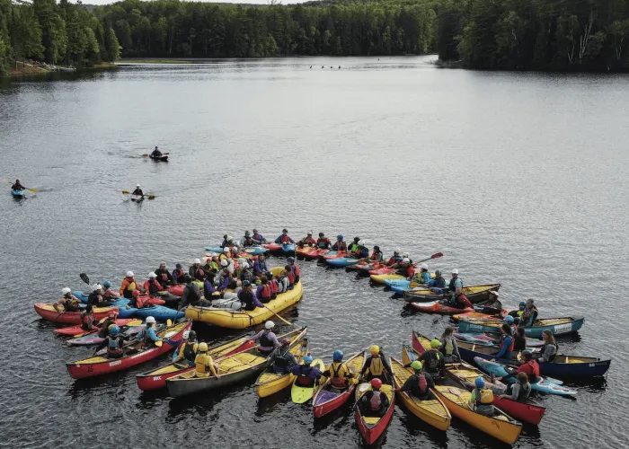 Several Kayaks grouped together in water around a raft, lake and forest in backgroun